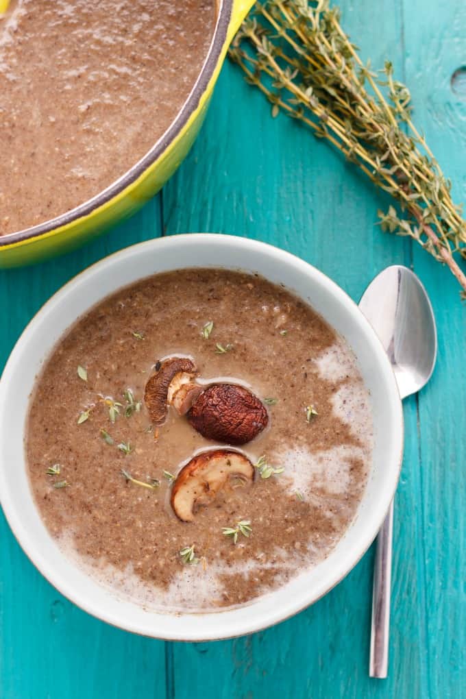 Milestones' Roasted Mushroom Soup in white bowl and green pot on blue table with spoon and herb