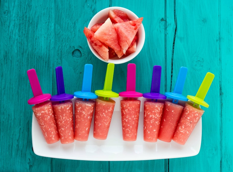 Watermelon and coconut popsicles on white plate near sliced watermelon in white bowl on blue table