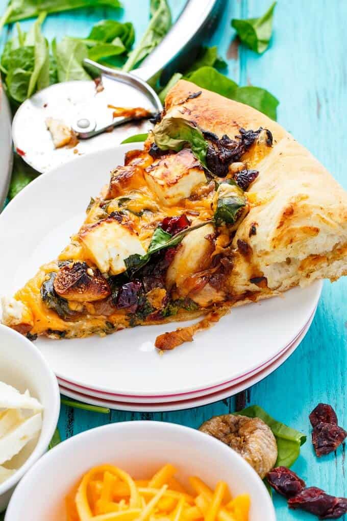 Caramelized Onion Dried Fig Pizza with Feta slice on small white plate next to bowls and pizza cutter and veggies on blue table