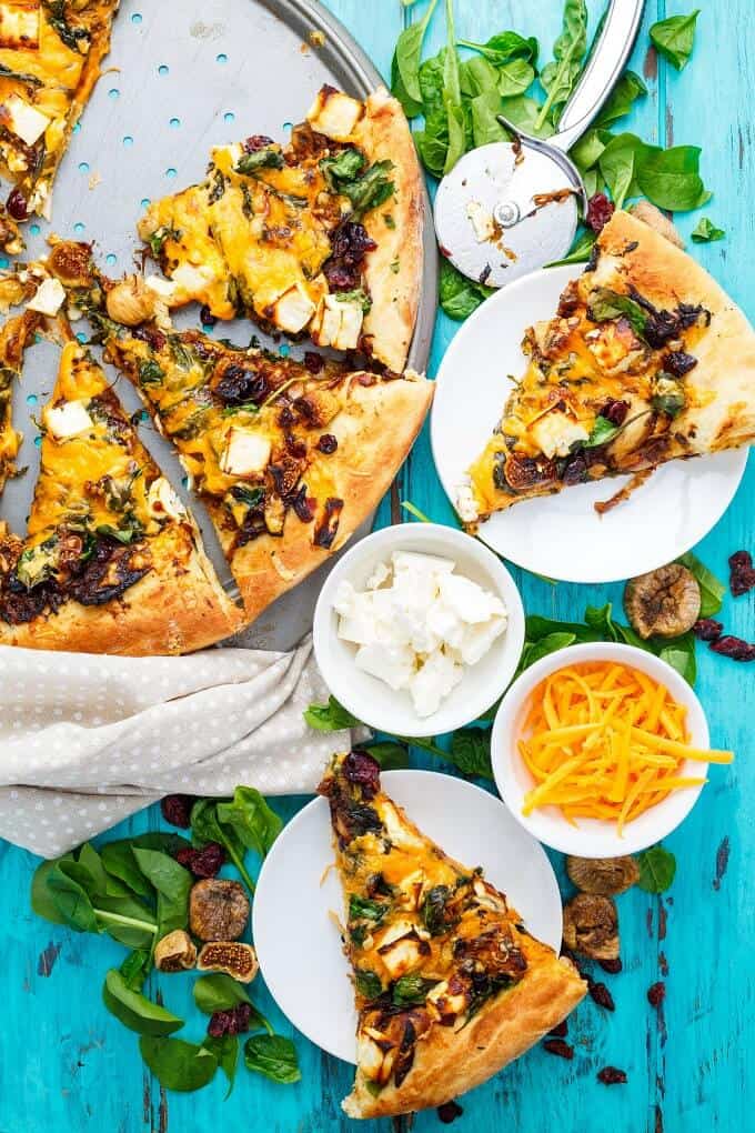 Caramelized Onion Dried Fig Pizza with Feta on gray tray and white plates. Pizza cutter, herbs, bowls of ingredients on blue table