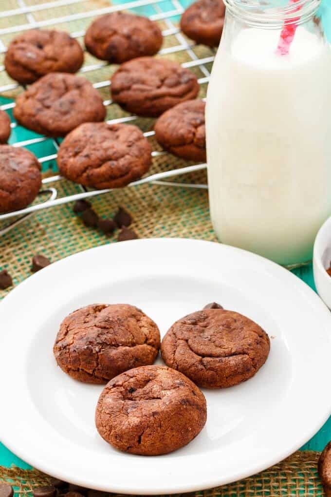 Double Chocolate Cookies with Hershey's Chipits on white plate next to glass jar full of milk and cookies on baking grid on table with chocolate chips