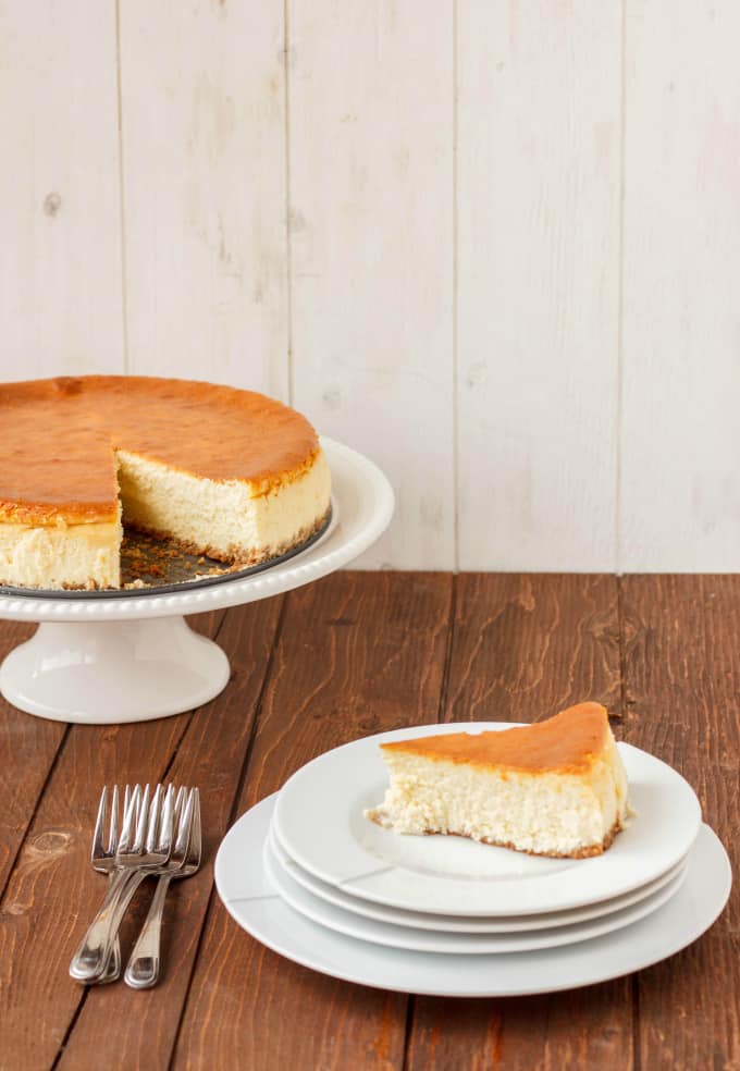 New York Style Cheesecake with Strawberry Topping on white plate on wooden table next to forks and stray with cheesecake