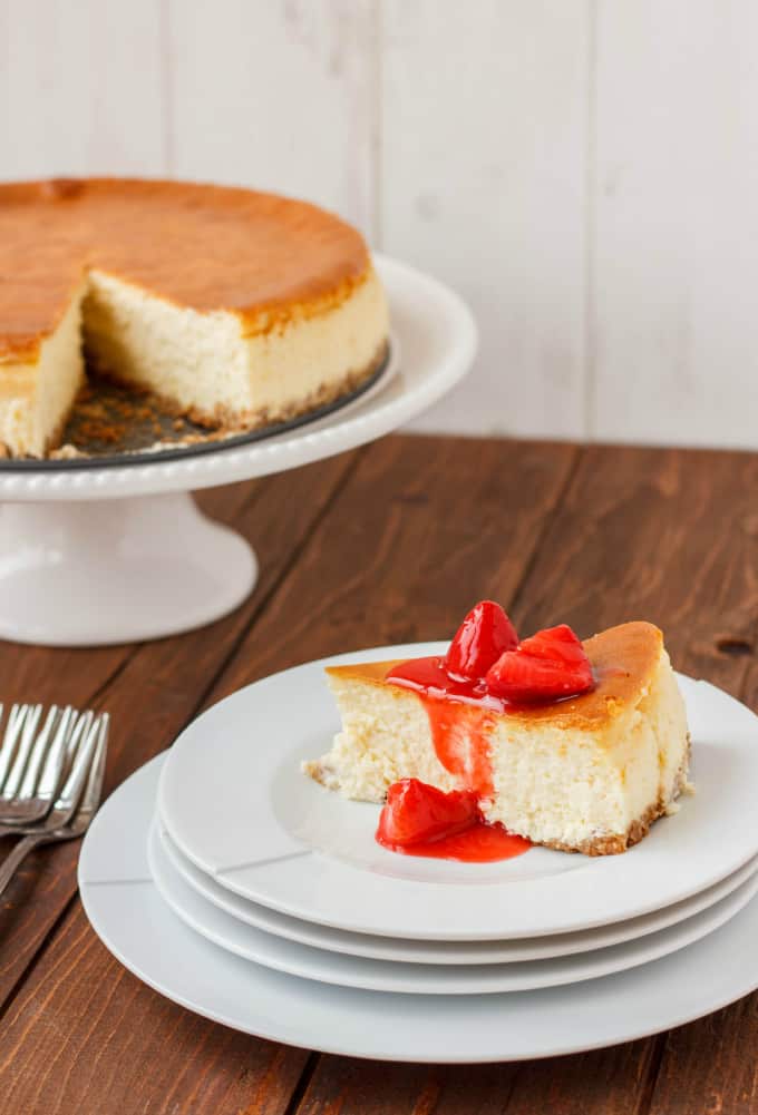 New York Style Cheesecake with Strawberry Topping on white plates next to forks and cheesecake on white stray on wooden table