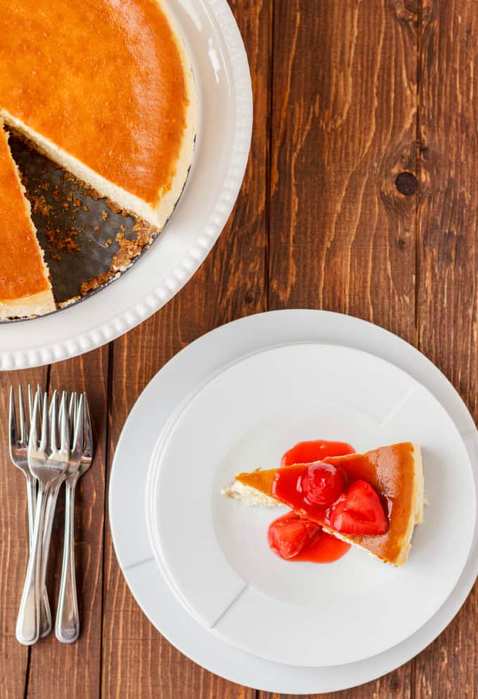 New York Style Cheesecake with Strawberry Topping on white plate on wooden table next to forks and cheesecake on white stray,