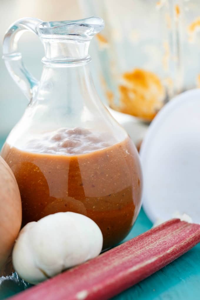 Rhubarb BBQ Sauce  in glass jar with rhubard, garlic and onion on blue table