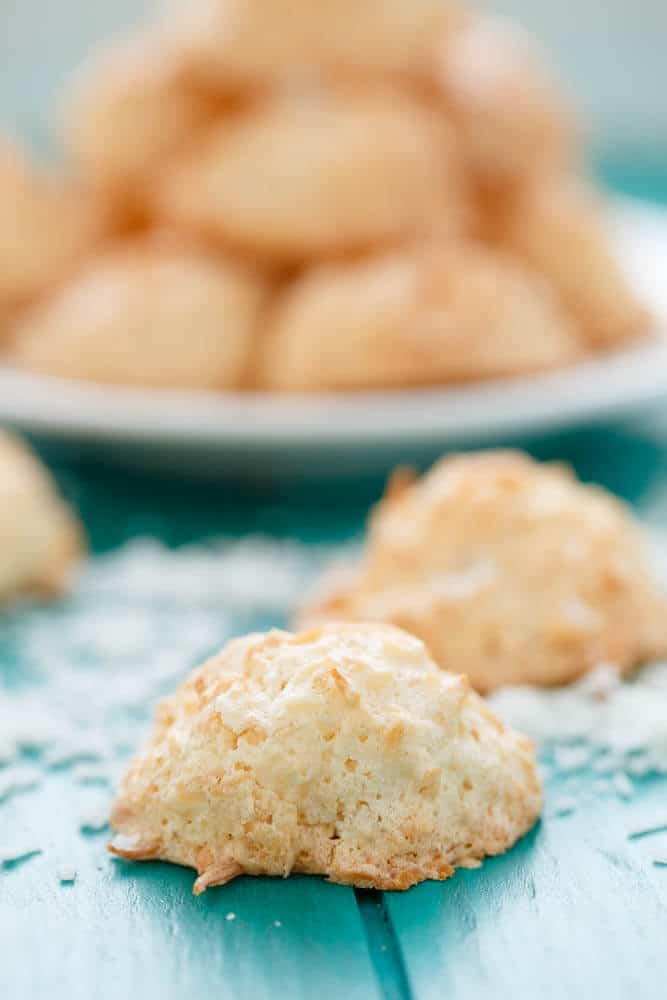 Semi-Toasted Coconut Macaroons on blue table sprinkled by sliced coconut, plate with cakes in the background#glutenfree