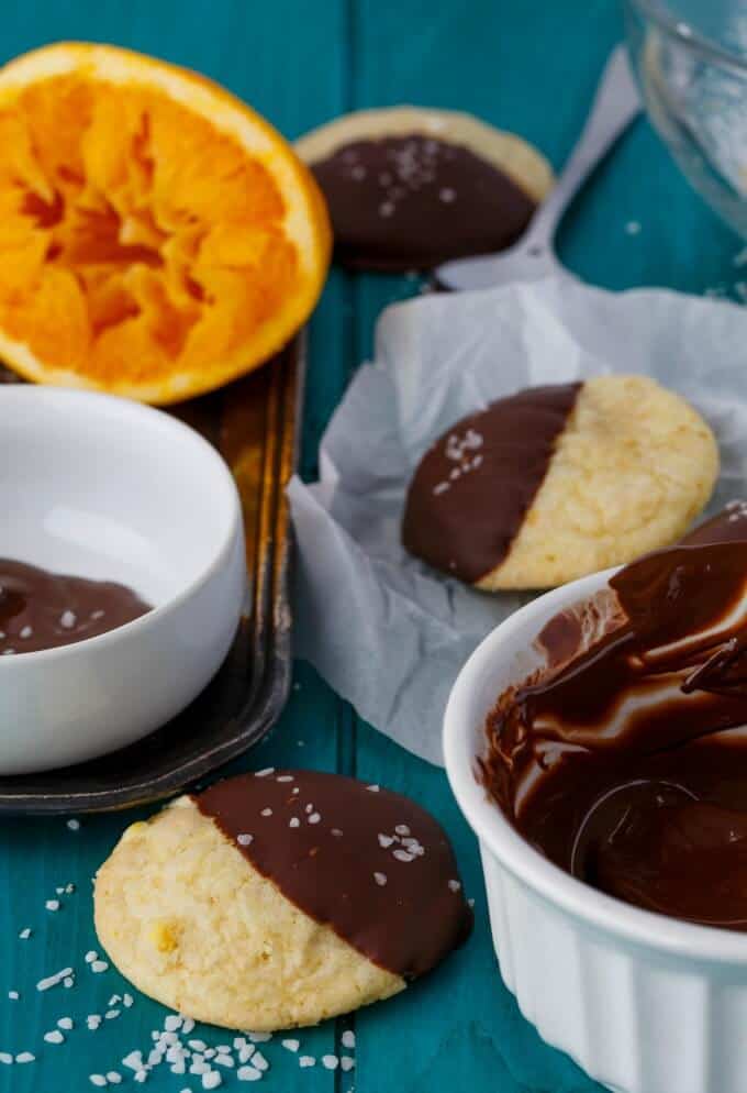 Chocolate Dipped Orange Cookie Crinkles on blue table next to sliced oranges, spoon, chocolate dip in bowls