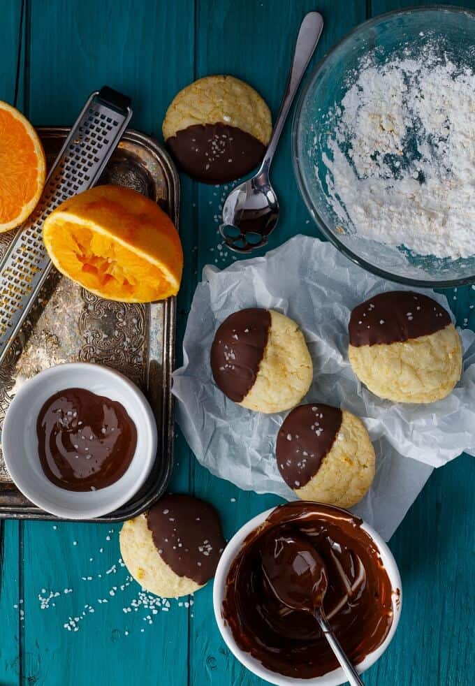 Chocolate Dipped Orange Cookie Crinkles on blue table next to sliced oranges, bowls of chocolate dip, spoon and bowl of flour