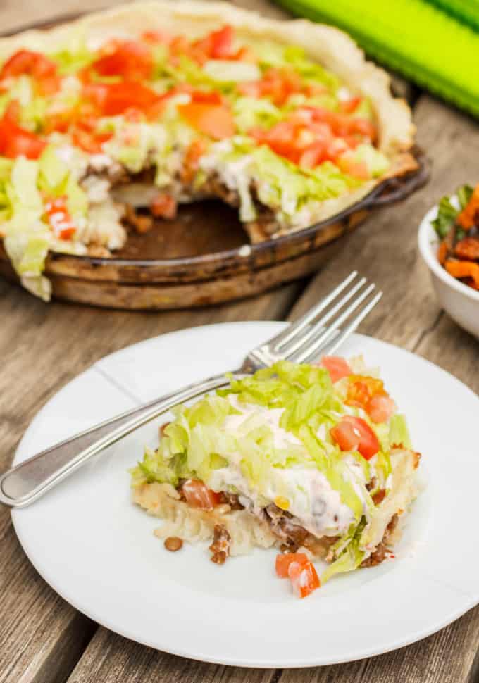 Gluten-Free and Vegan Taco Pie on white plate with fork on wooden table, in background taco pie on big glass plate