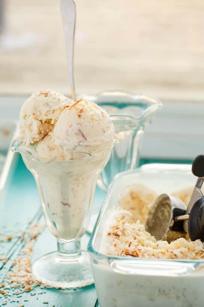 Toasted Coconut Ice Cream in tall glass with spoon on blue table next to empty glass and glass jar full of icecream with icecream spoon#coconut