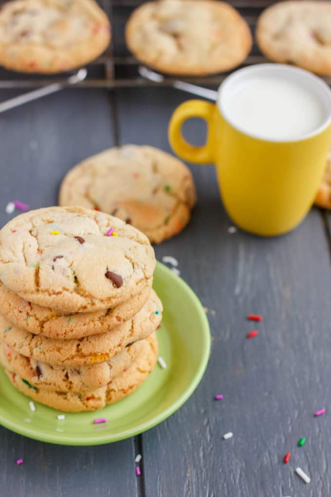 Birthday Cake Chocolate Chip Cookies on small green plate on black table with cookies in the background and yellow mug with milk