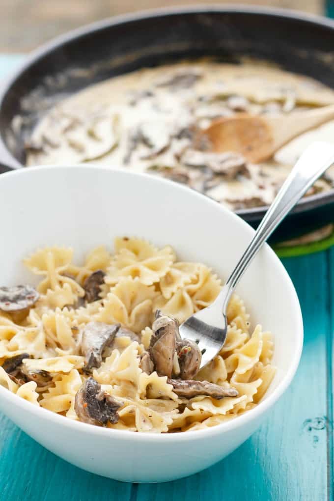 Portobello Mushroom Pasta with Cream Sauce in white bowl with fork on blue table, sauce in pan in the background#glutenfree