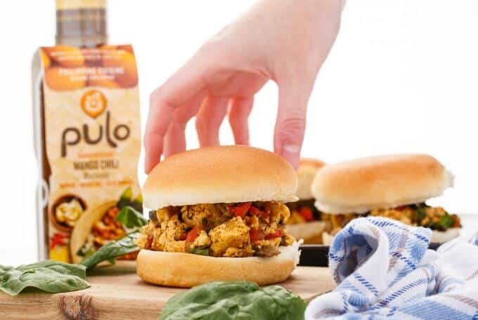 Pulo Mango Chili Vegan Sloppy Joes burger held by hand on wooden kitchen pad, pulo marinade in the background
