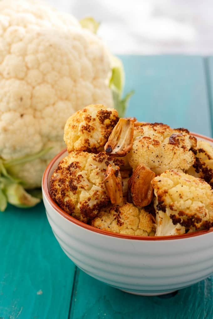 Roasted Cauliflower with Garlic in white bowl on blue table with fresh cauliflower in the background#vegan #garlic