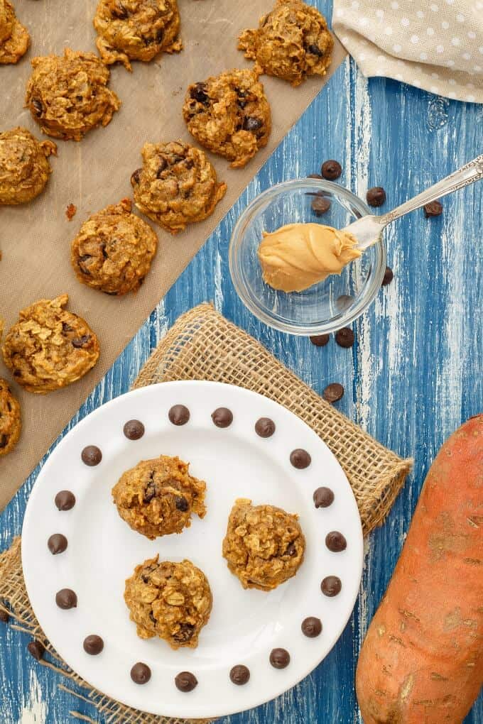 Sweet Potato Chocolate Chip Cookies on white plate on table next to glass bowl with peanut butter and knife and cookies on baking paper and sweet potato