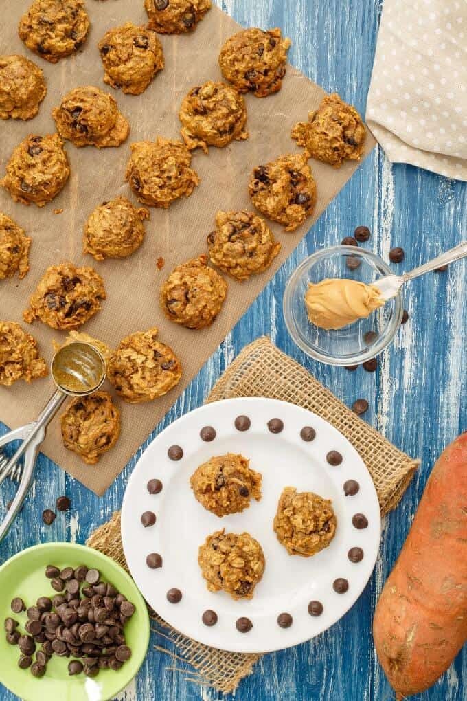 Sweet Potato Chocolate Chip Cookies on white pare next to sweet potato, glass bowl of peanut butter on knife, green bowl of chocolate chips and cookies on sheet of baking paper
