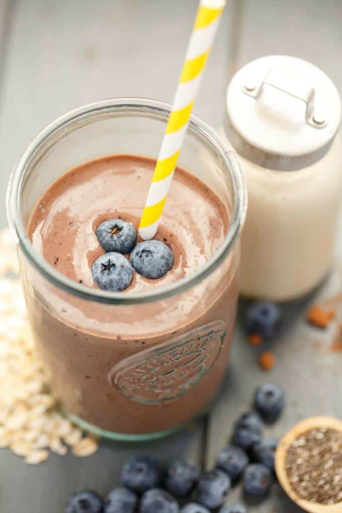 Almond Milk Banana-Blueberry Breakfast Smoothie in glass with starw and blueberries on the top next to jar of milk on gray table with blueberries and oats #vegan