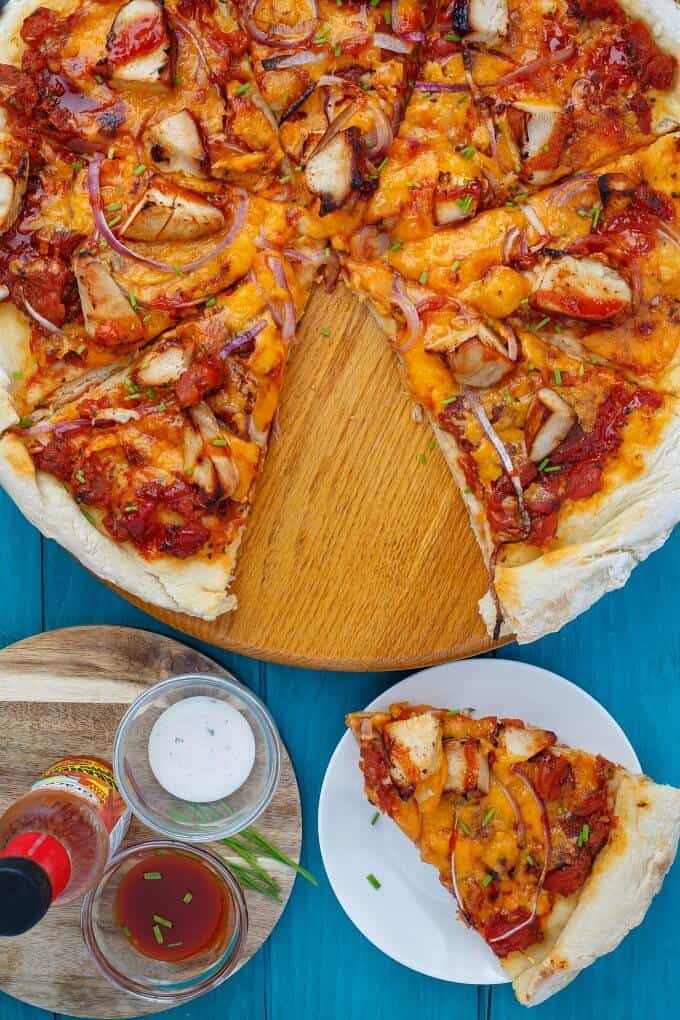 BBQ Chicken Pizza with Mushrooms and Onionson wooden pad and white plate next to spicy sauce in bowls and bottle on blue table