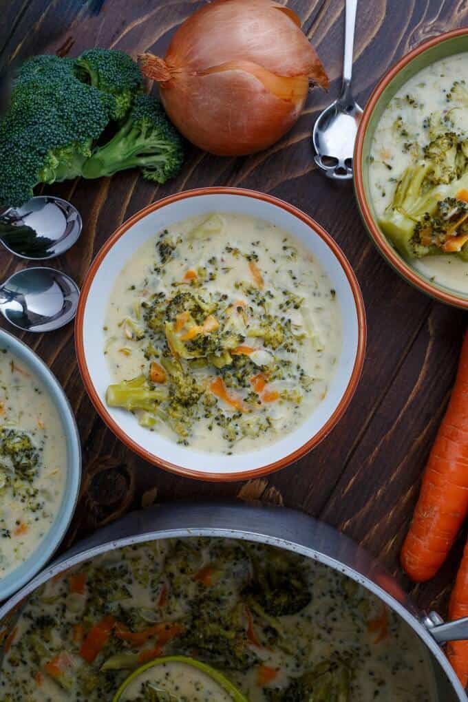 Cream of Broccoli Soup in bowl, pot and plate on brown table with spoons, carrots and broccoli