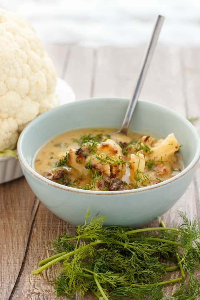 Vegan Roasted Cauliflower Soup in white bowl with spoon on brown table with veggies#almondbreeze