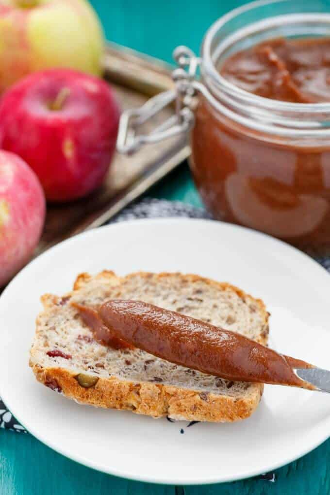 Apple Butter in the Slow Cooker in glass jar next to apples, white palte with slice of bread and knife with apple butter on blue table