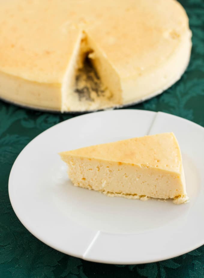 Crust-Less Lemon Cheesecake slice on white plare on blue table, rest of cheesecake in the background