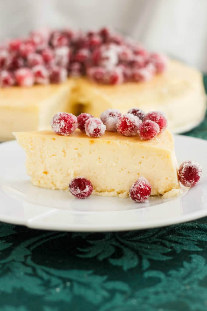 Crust-Less Lemon Cheesecake with Candied Cranberries slice on white plate on blueish table, rest of cheesecake in the background