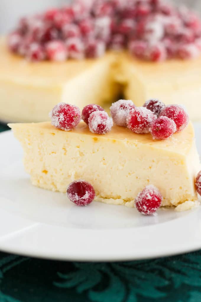 Crust-Less Lemon Cheesecake with Candied Cranberries on white plate, rest of the cake in the background