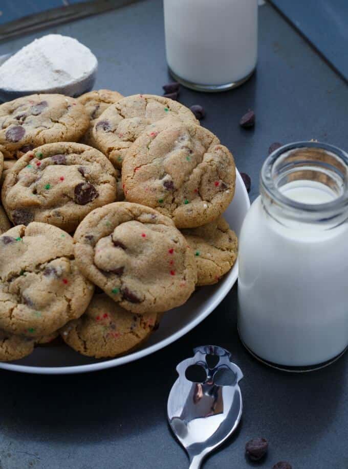 Gluten-Free Peppermint Cookies with Chocolate Chips on white plate on black table next to glass jars of milk, spoon, chocolate chips and flour in bowl