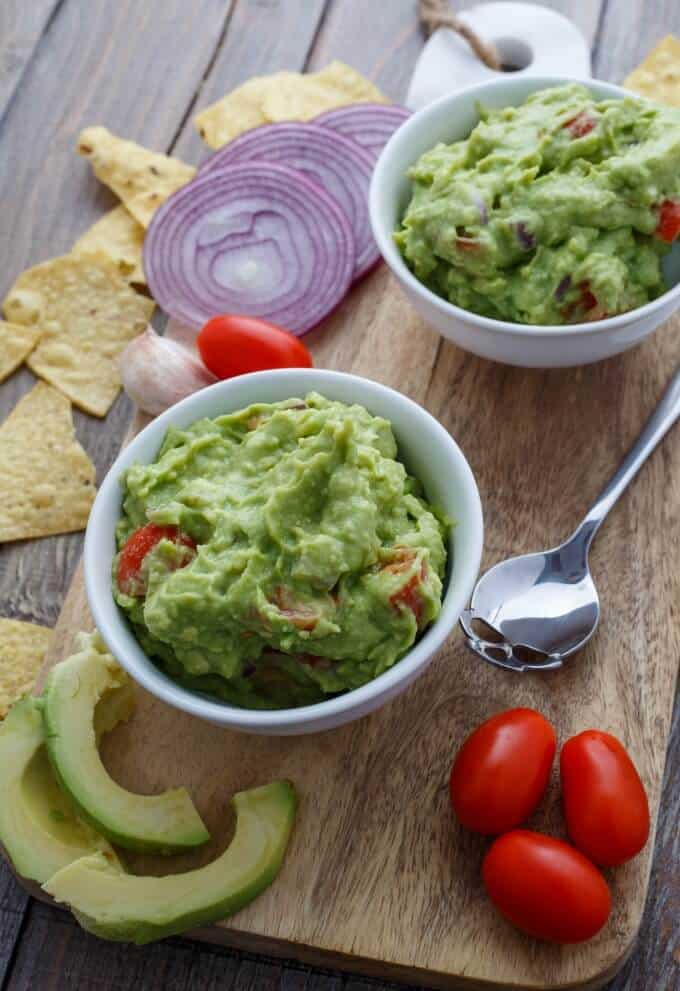 Homemade Guacamole Dip in white bowl on wooden pad, on wooden table with spoon, tomatoes, sliced guacamola, sliced onions, garlic and chips