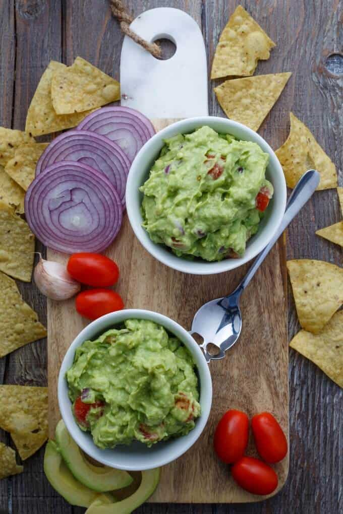 Homemade Guacamole Dip in white bowls on wooden pad on wooden table with spoon, sliced guacamola, sliced onion, tomatoes, garlic and chips