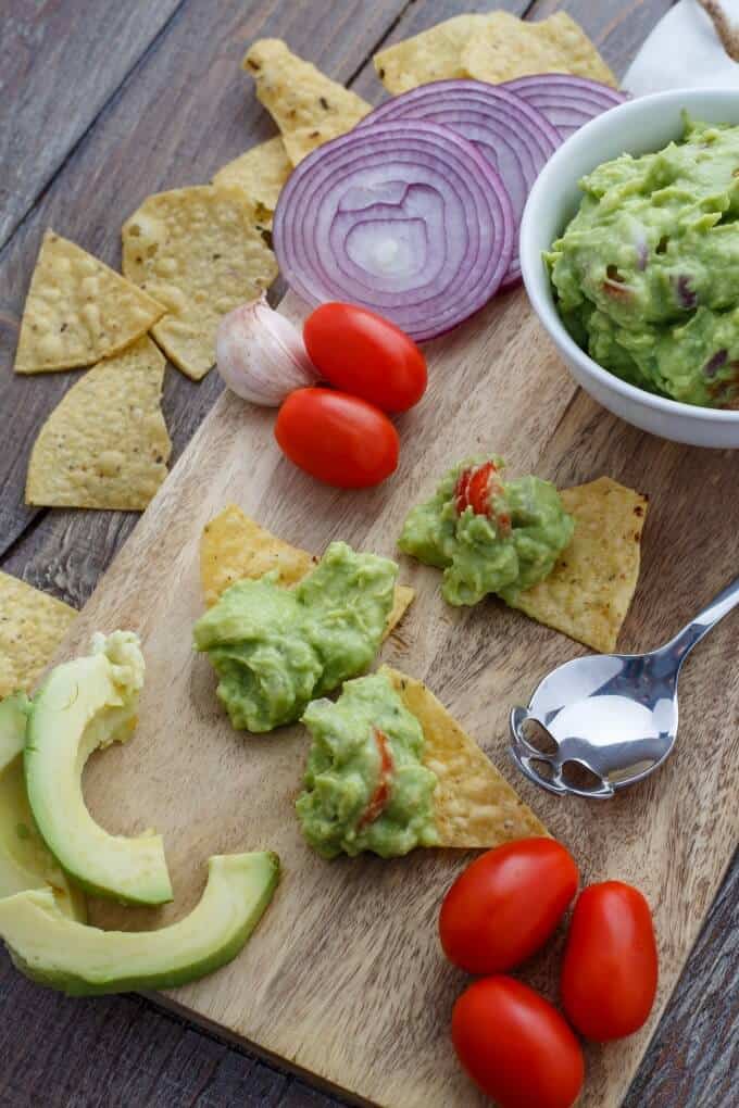 Homemade Guacamole Dip in white bowl on wooden pad on wooden table with spoon, guacamola slices, sliced onion, tomatoes, garlic and chips with and without dip