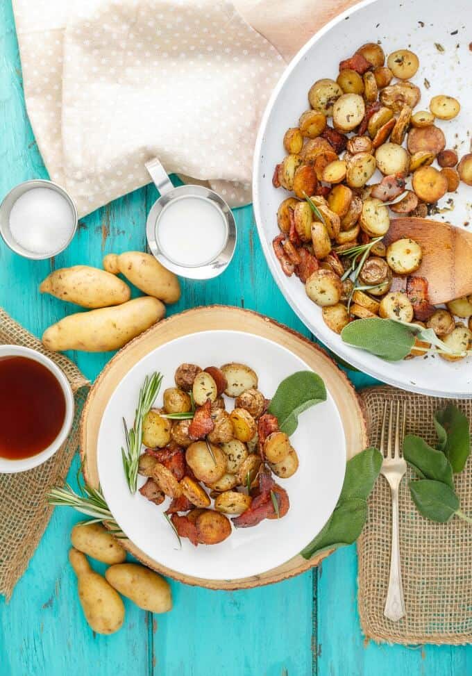 Pan-Fried Fingerling Potatoes with Bacon on white plate and pan with saptula on blue table with forsk, potatoes, bowl of sauce, milk, slat and veggie around