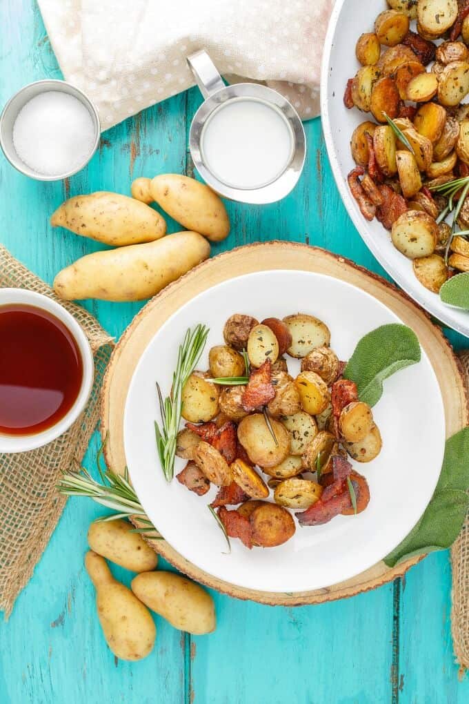 Pan-Fried Fingerling Potatoes with Bacon on white plate and in white pan with herbs. Potatoes, bowl of sauce, jar of milk and bowl of ingredient on blue table