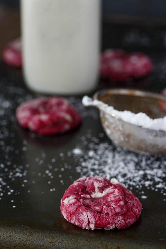 Red Velvet Cookie Crinkles on black table next to sprinkler and bottle of milk