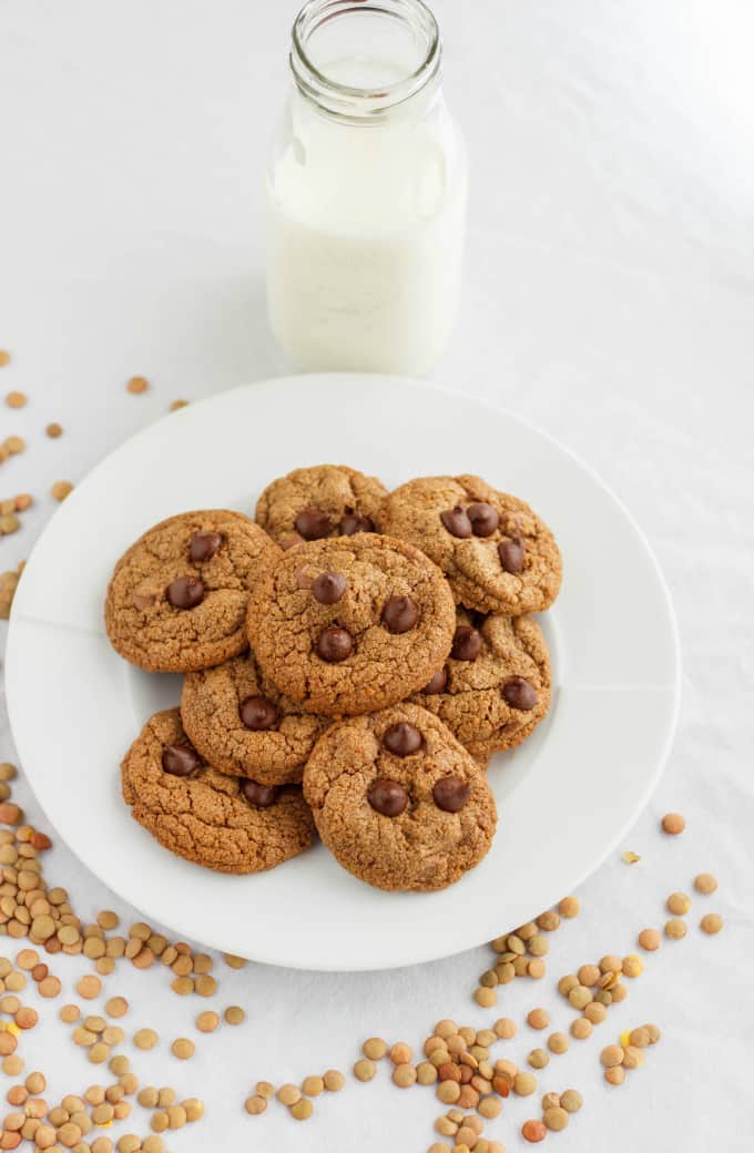 Chocolate Chip Cookies made with Lentil Flour on white plate and white table, lentil poured around plate and glass bottle of milk