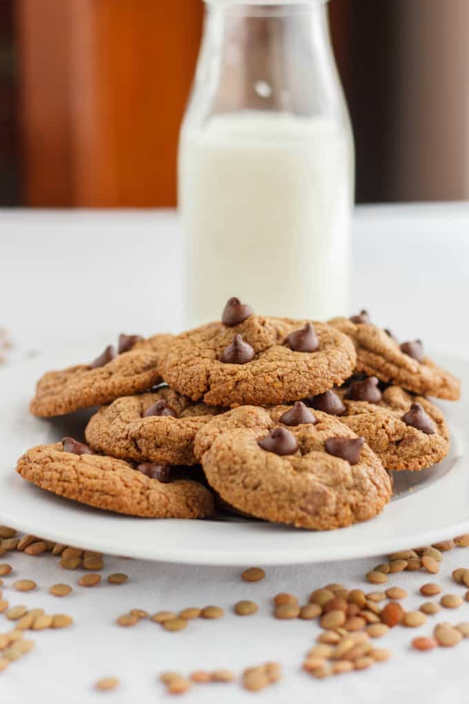 Chocolate Chip Cookies made with Lentil Flour on white plate, lentils around plate on white table, in the background glass bottle of milk