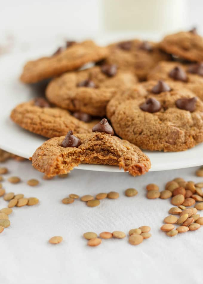 Chocolate Chip Cookies made with Lentil Flour on white plate, one is half eaten, lentils around plate on white table