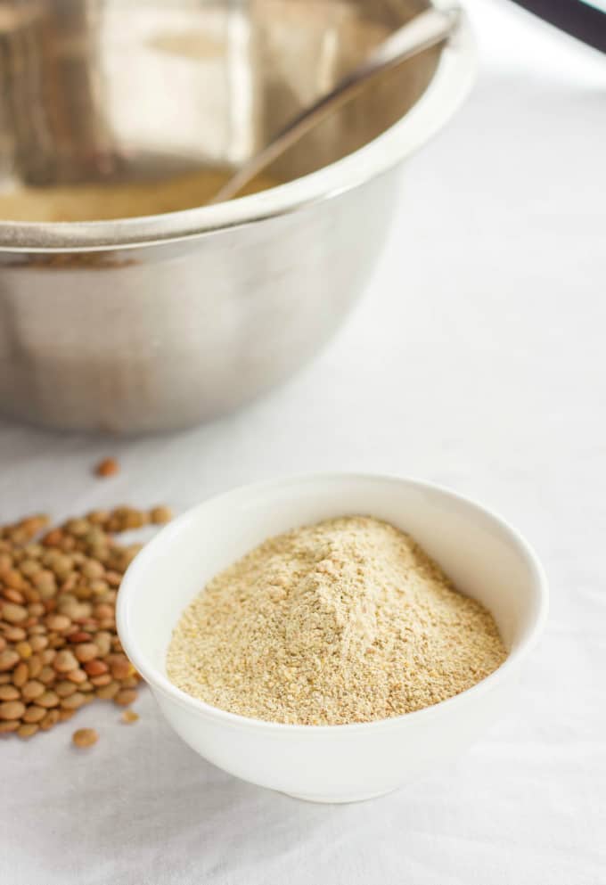 Homemade Lentil Flour  in white bowl next to big bowl with scoop on white table with spilled lentils