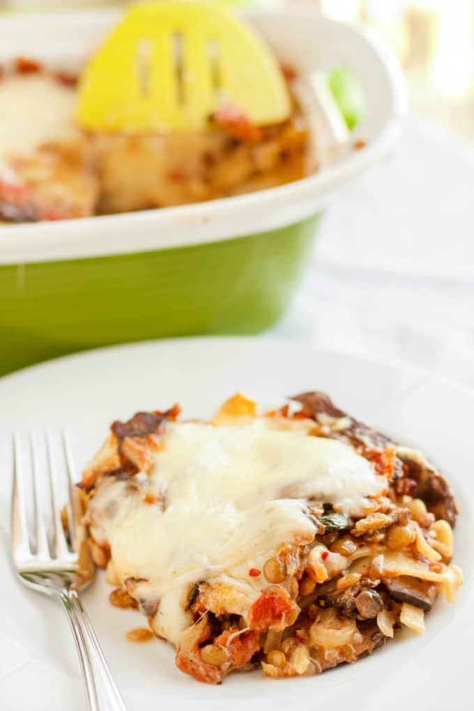 Lentil lasagna in roasted portobello cream sauce on white plate with fork, Green pot with same dish in the background