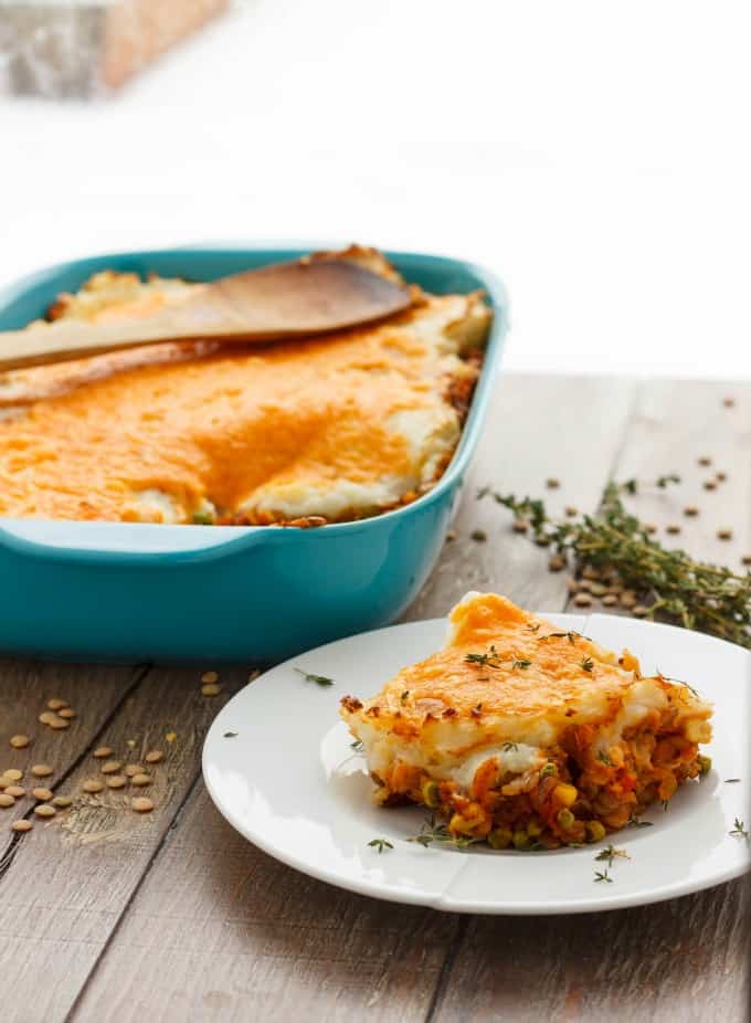 Vegetarian Shepherd's Pie slice on white plate on wooden table next to blue baking pot full of pie with wooden spatula (Vegan Shepherd's Pie Included.) on #vegetarian