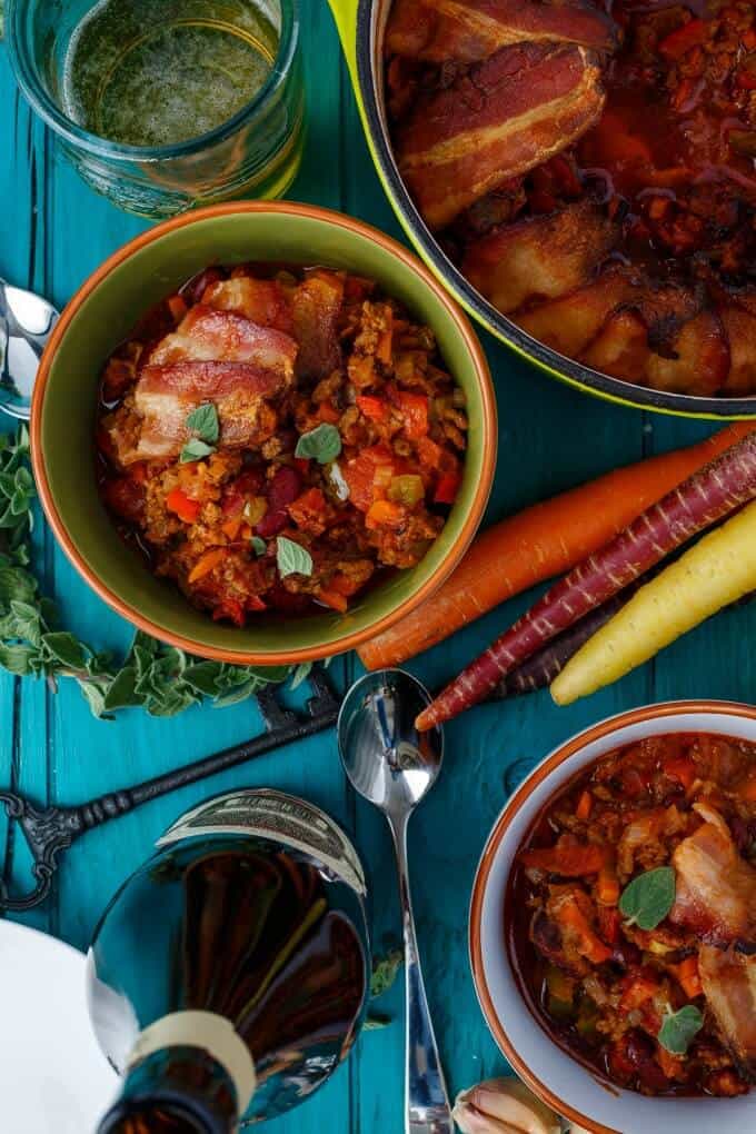 Beer and Beef Chili in a Bacon Bowl on blue table with pots,key, spoon, beer botle and variety of vegetables