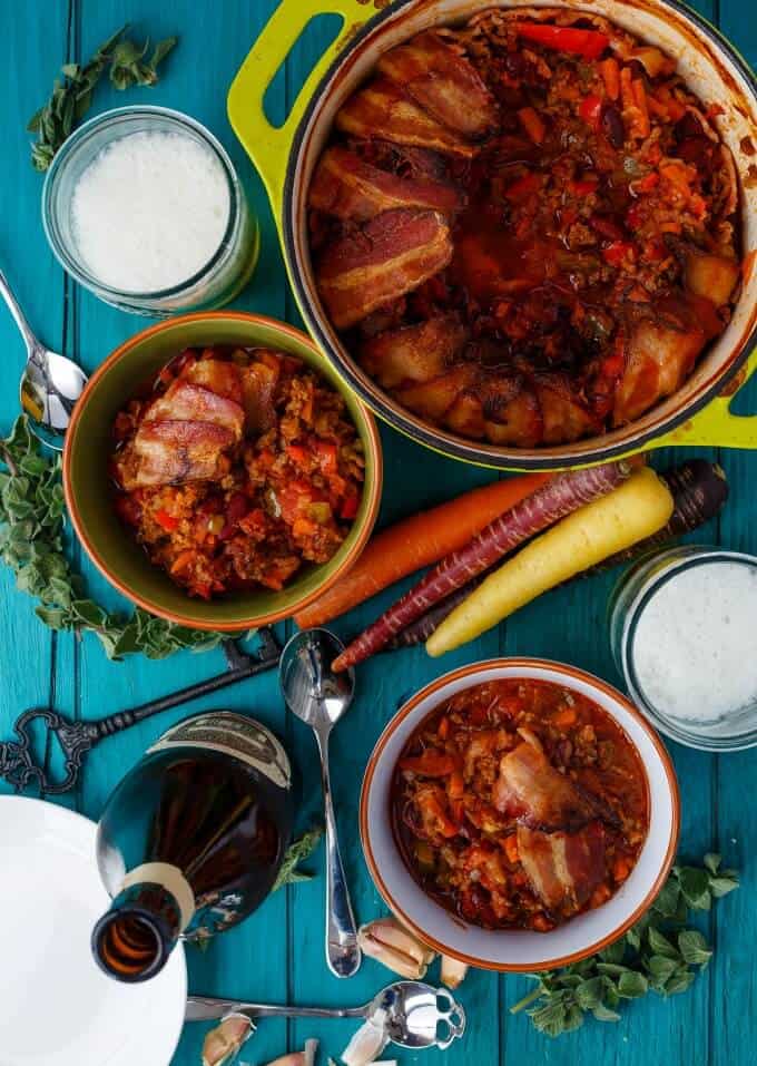 Beer and Beef Chili in a Bacon Bowl in bowls and pot. Key, bottle beer, vegetables, herbs, white plate, spoon and ingredients in cups on blue table