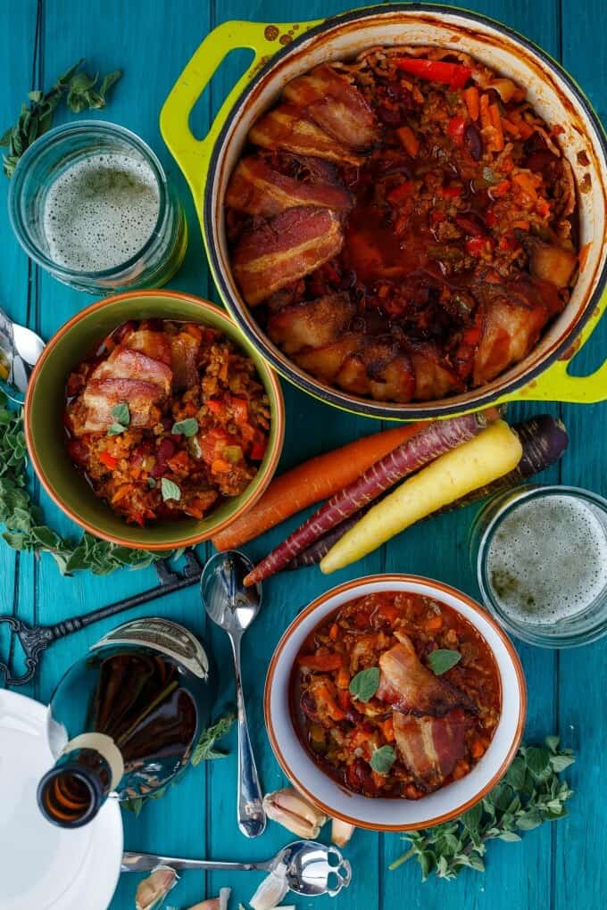 Beer and Beef Chili in a Bacon Bowl on blue table with pots,, key,spoon,beer bottle, and many vegetables