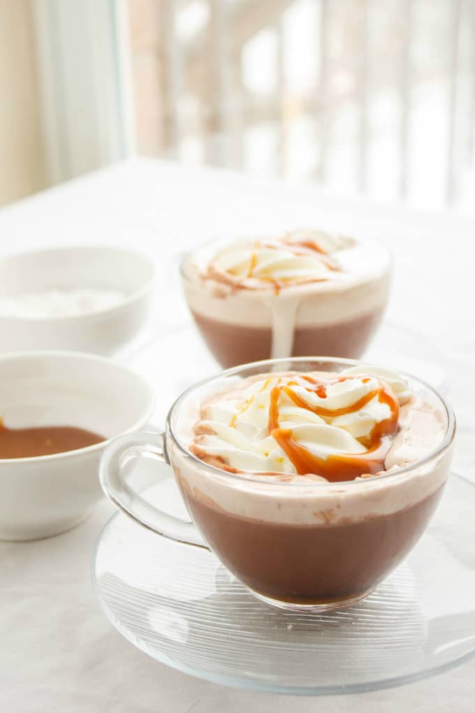 Salted Caramel Hot Cocoa in glass cups on glass plates next to white bowls on white table