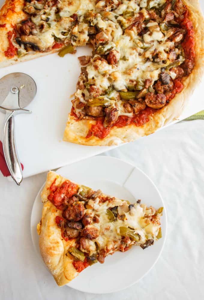 Sausage Pizza with Onions, Green Peppers, and Bacon on white tray with pizza cutter, slice of pizza on white plate
