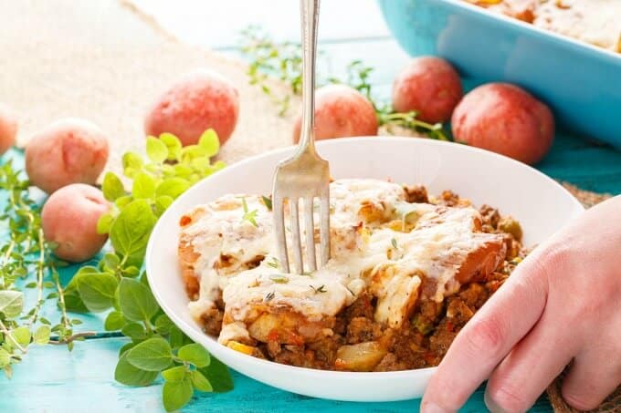 Smashed Potato Shepherd's Pie on white placked with fork on table with hand, potatoes,veggies and baking pot(Cottage Pie)