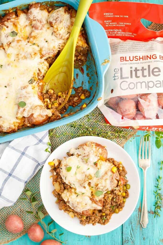 Smashed Potato Shepherd's Pie  in blue casserole with green spatula, on white plate. Fork, herbs, potaotes, sack of potatoes and cloth wipe on blue table