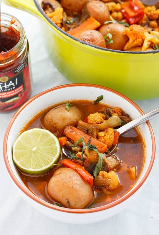 Roasted Vegetable Massaman Curry Soup in orange white bowl with spoon on white table, green pot with same dish and glass jar in the background(Coconut-Free) #vegan