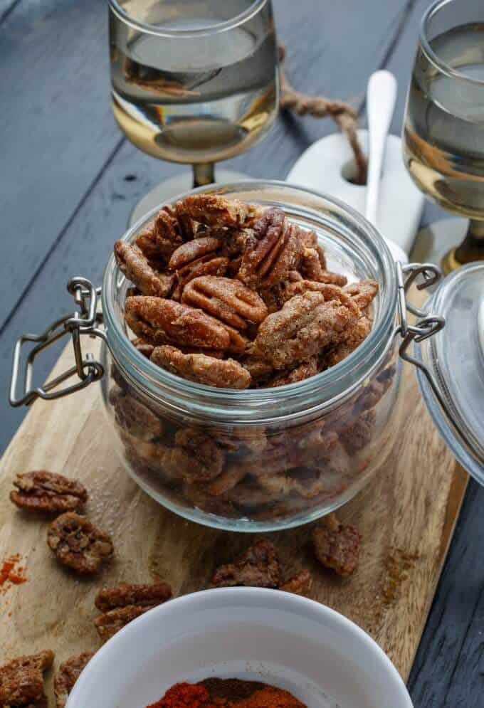 Spicy Glazed Pecans in glass jar on wooden pad next to glasses of wines and nuts